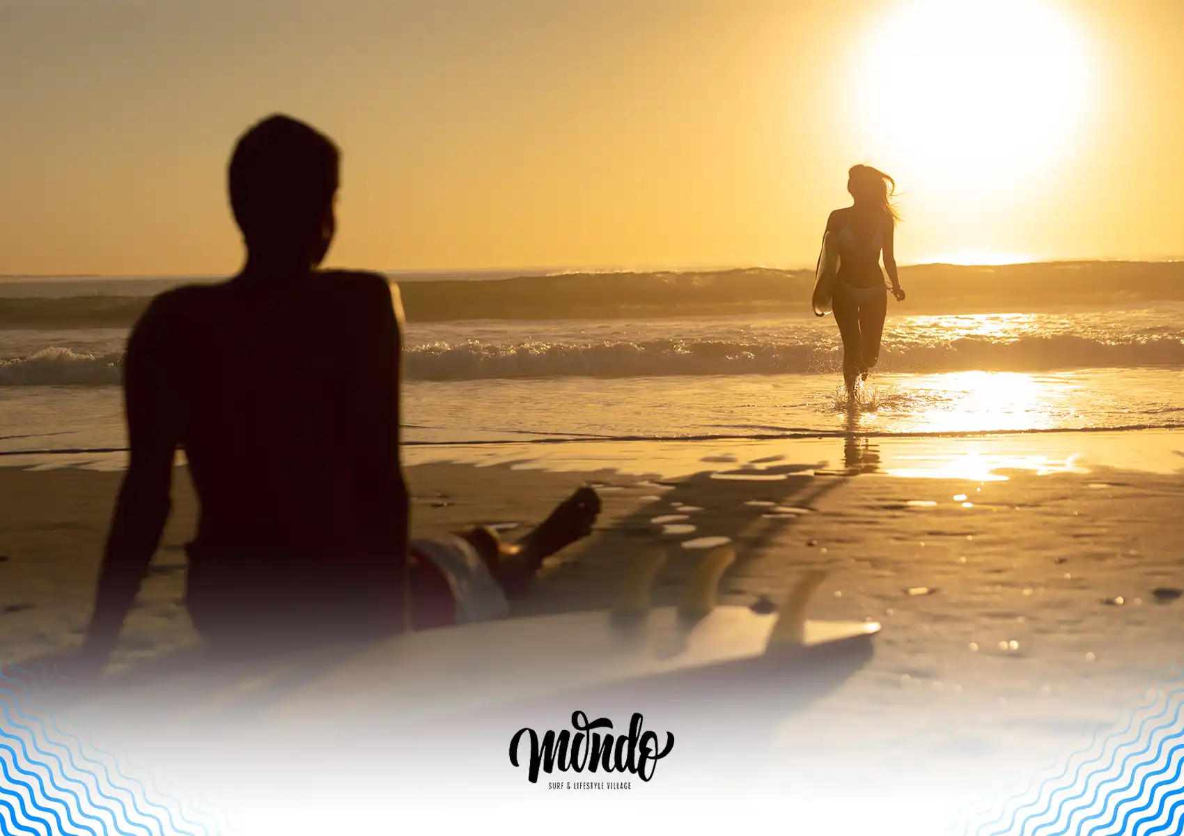 A surfer sits resting on the sandy beach in the foreground, watching another surfer carry her board out of the sparkling ocean waves into a brilliant golden sunrise at a surf camp bali.