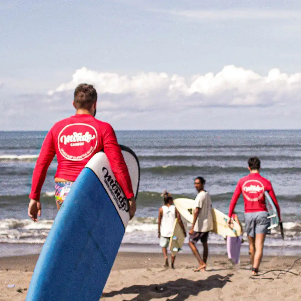 A group of surfers carrying their boards across a sandy beach towards the ocean waves during a session at a surf camp Bali, with the person in the foreground wearing a red rash guard and holding a large blue surfboard.