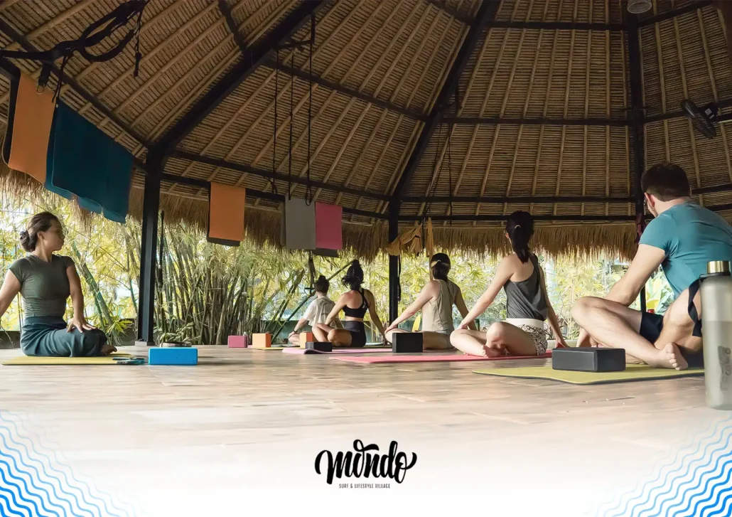A group of students practicing seated twists on yoga mats inside an open-air bamboo studio at a surf camp, Bali.