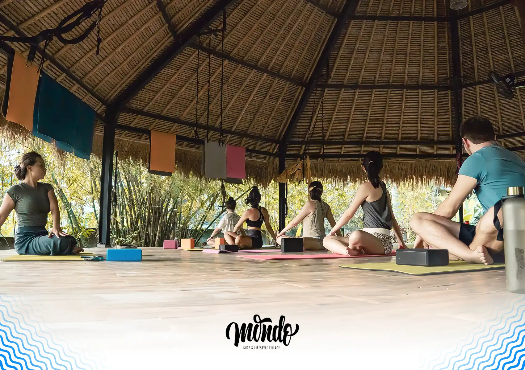 A group of students practicing seated twists on yoga mats inside an open-air bamboo studio at a surf camp, Bali.