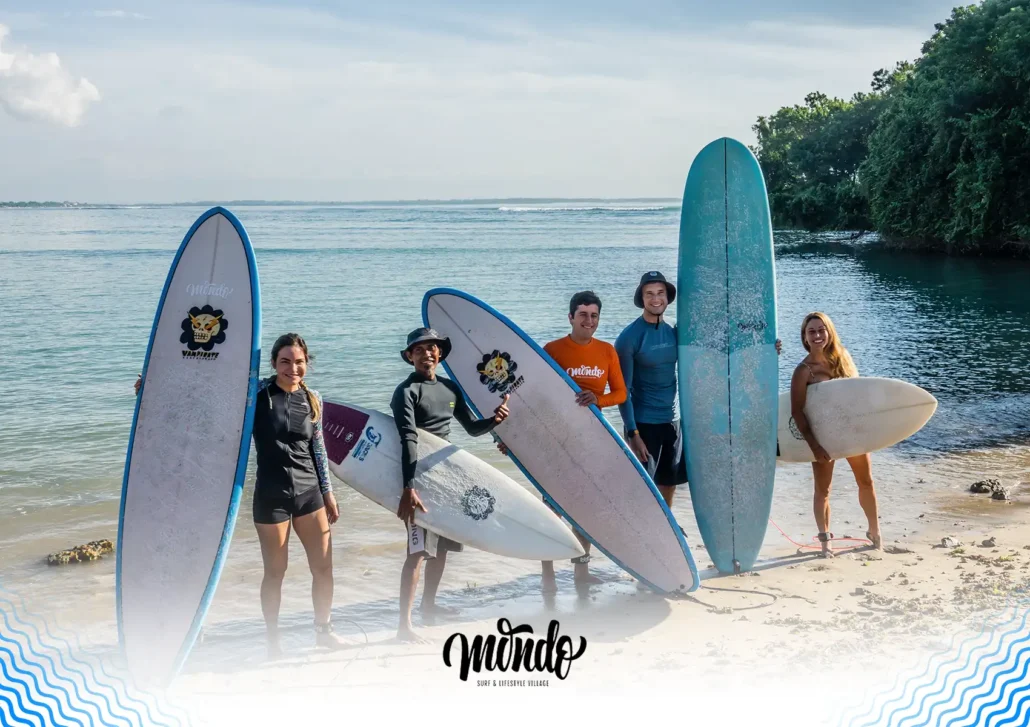 A group of smiling beginner surfers and a local instructor standing on the beach holding their surfboards at a surf camp, Bali.
