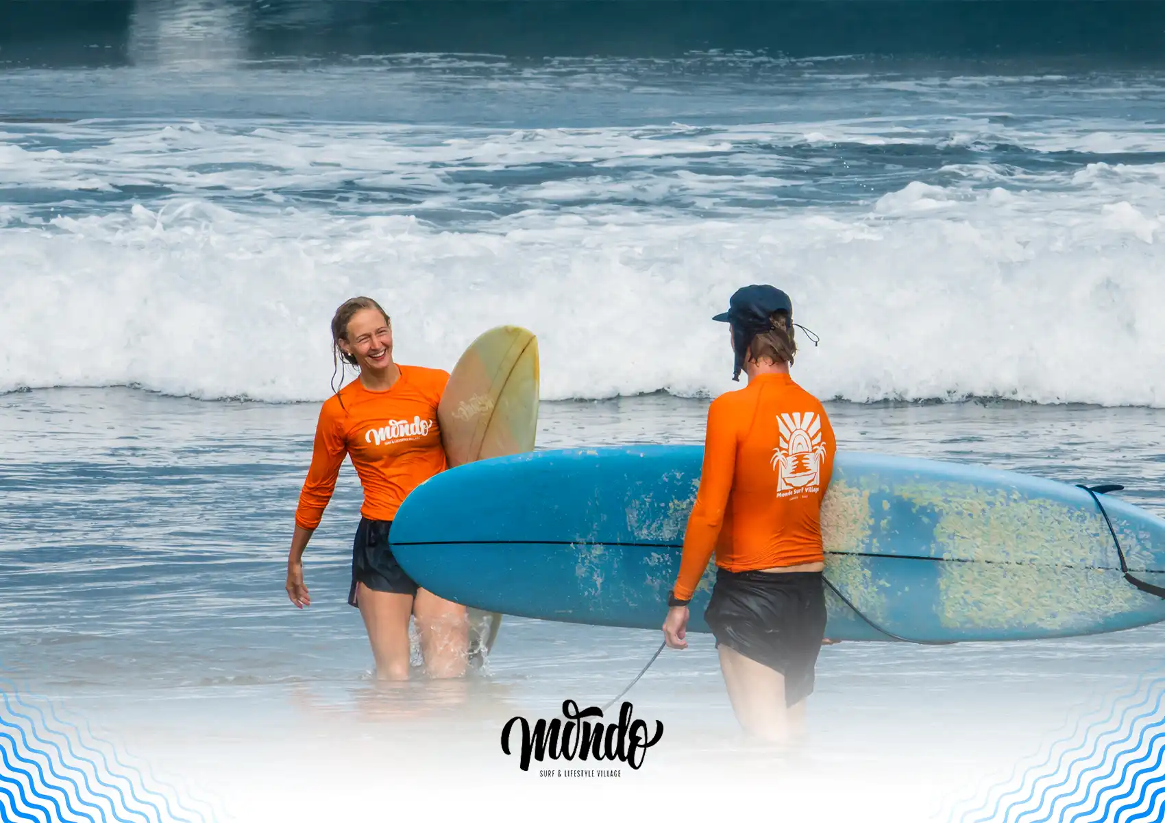 A smiling female student and her instructor, both wearing bright orange Mondo rash guards, wading through shallow white water with their surfboards during a lesson at a surf camp bali.