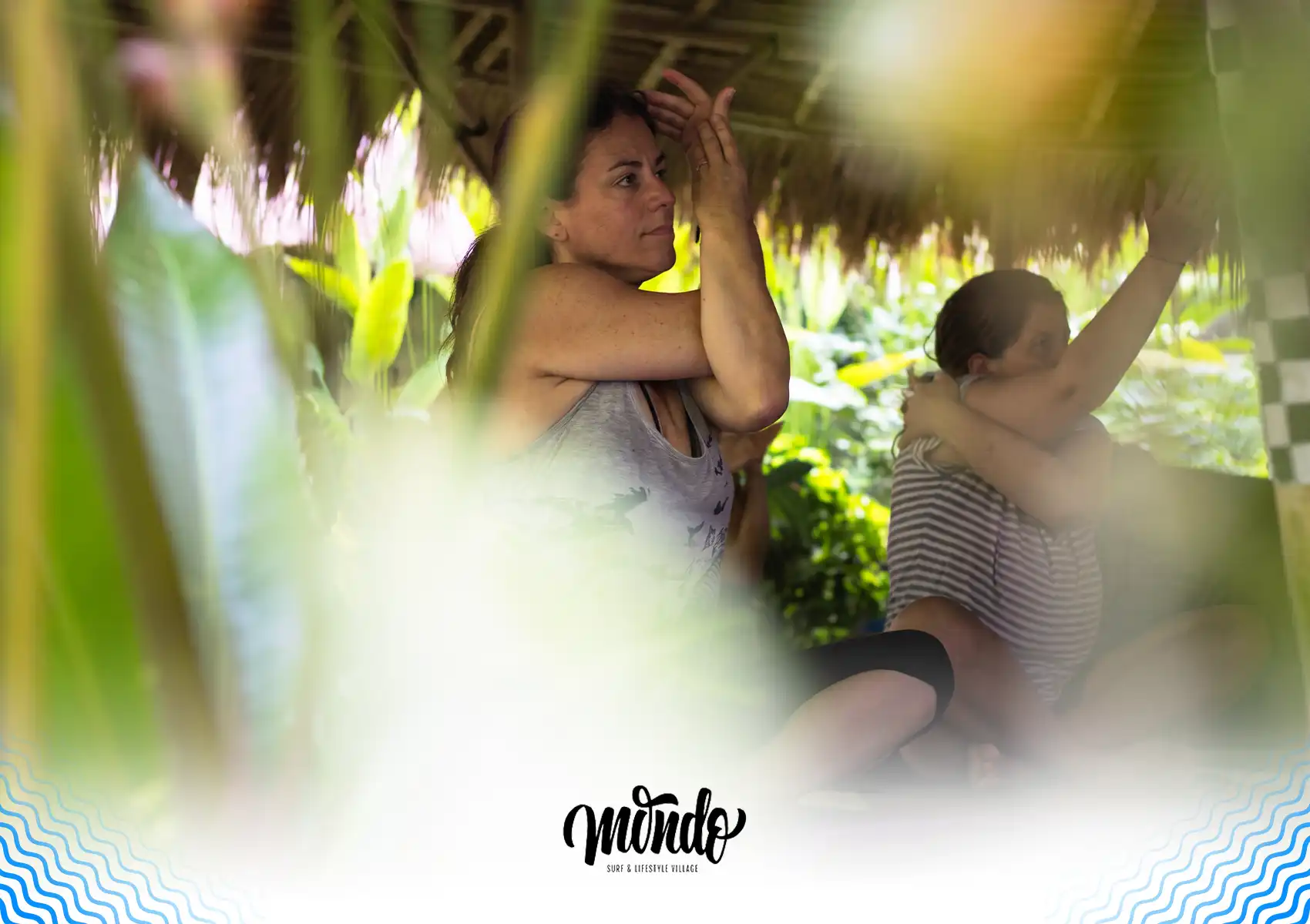 A woman practicing a seated arm stretch during a yoga session under a traditional thatched roof surrounded by tropical greenery at a surf camp, Bali.