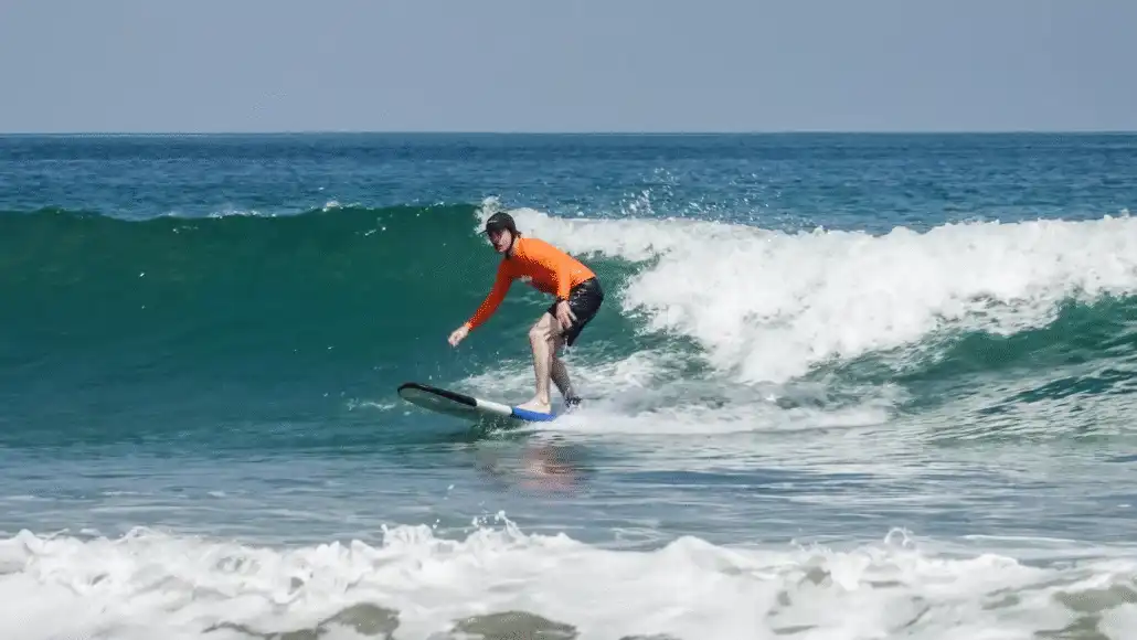 A surfer in an orange rash guard riding a wave on a soft-top surfboard during a session at a surf camp Bali.