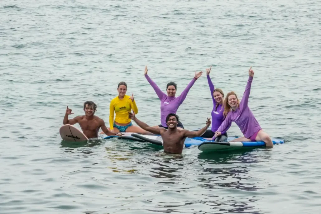 A group of friends and instructors cheering and throwing shaka signs on their surfboards in the ocean while attending a surf camp Bali.