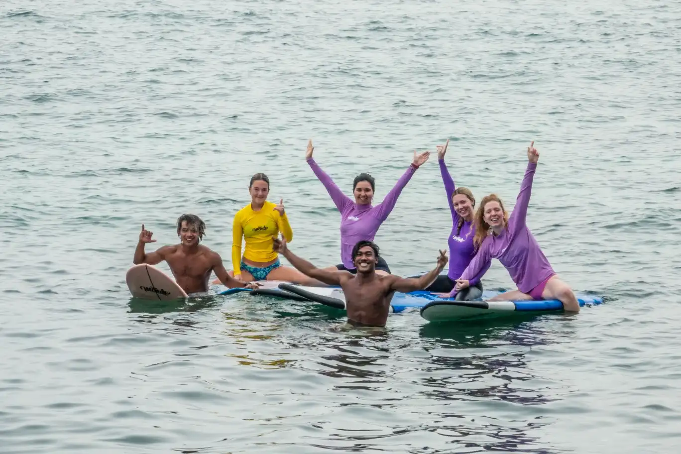 A group of friends and instructors cheering and throwing shaka signs on their surfboards in the ocean while attending a surf camp Bali.