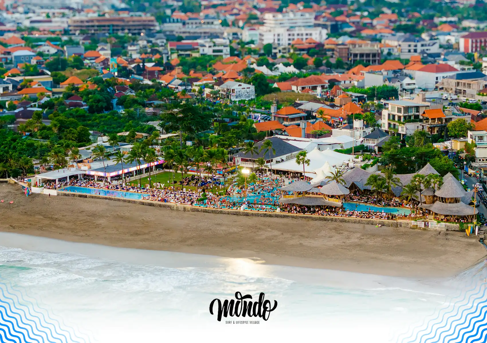 An expansive aerial view of a bustling beachfront club featuring large swimming pools, thatched-roof pavilions, and lively crowds right on the sand, highlighting the vibrant coastal lifestyle located near a surf camp bali.