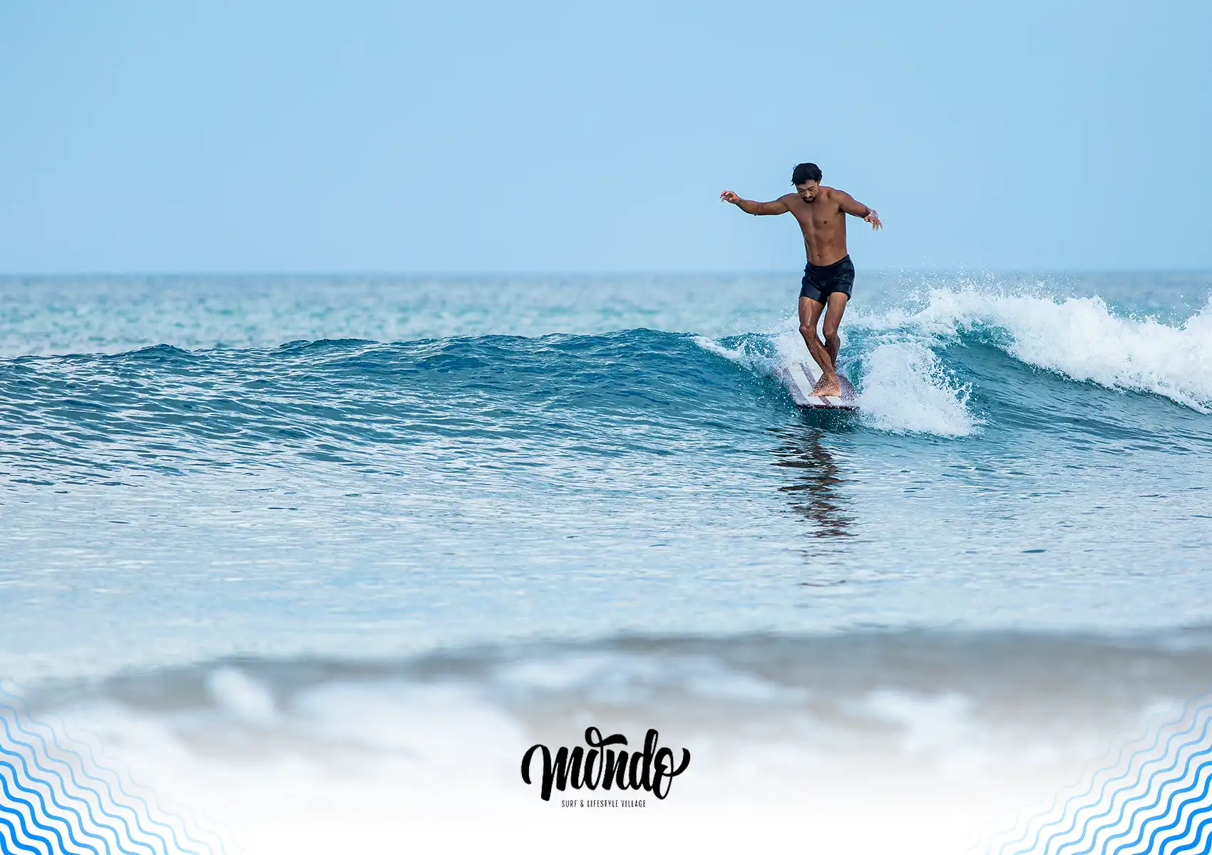 A skilled male surfer in black boardshorts confidently balances on his board while riding a smooth blue wave under a clear sky, illustrating the perfect surfing conditions you can enjoy while staying at a surf camp bali.