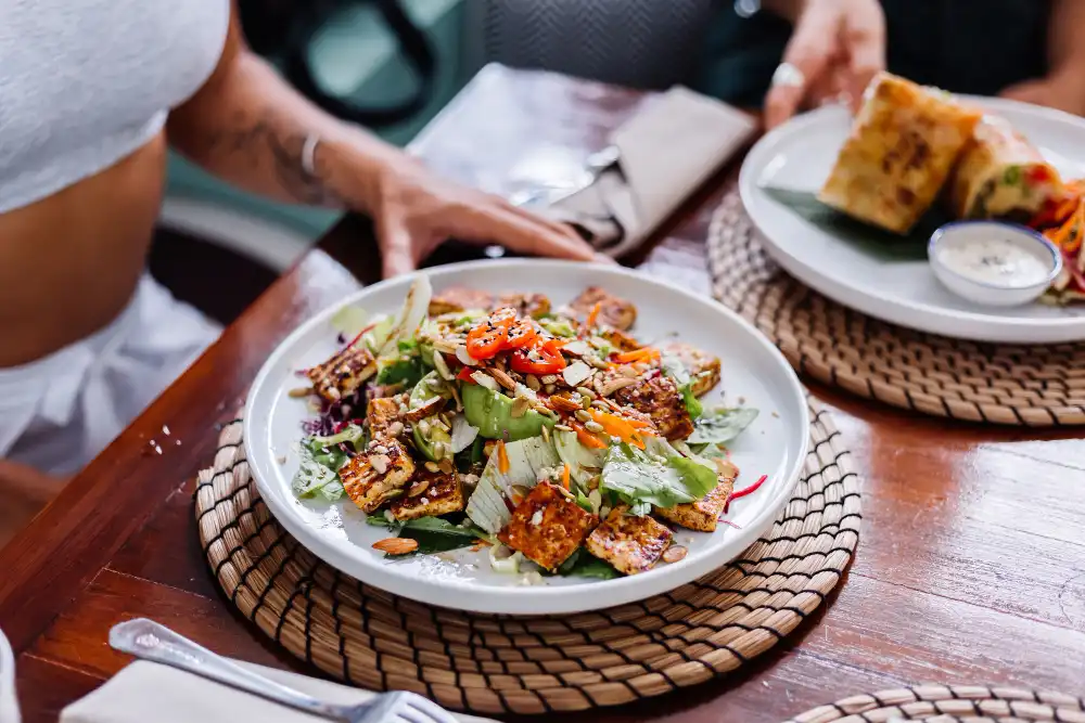 A fresh, healthy salad with grilled tofu, avocado, nuts, and seeds served on a woven placemat during lunch at a surf camp Bali, with a person sitting at the wooden table.
