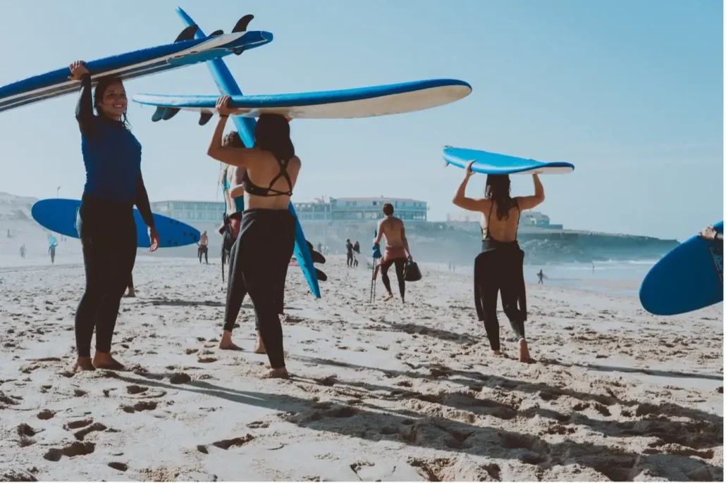 A group of surfers carrying long blue surfboards on their heads as they walk across a sunny beach during a surf camp Bali session.