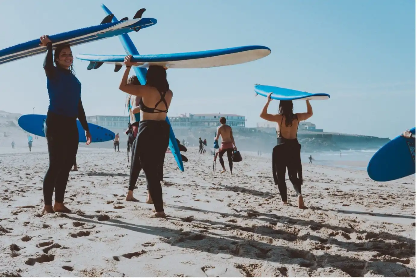 A group of surfers carrying long blue surfboards on their heads as they walk across a sunny beach during a surf camp Bali session.