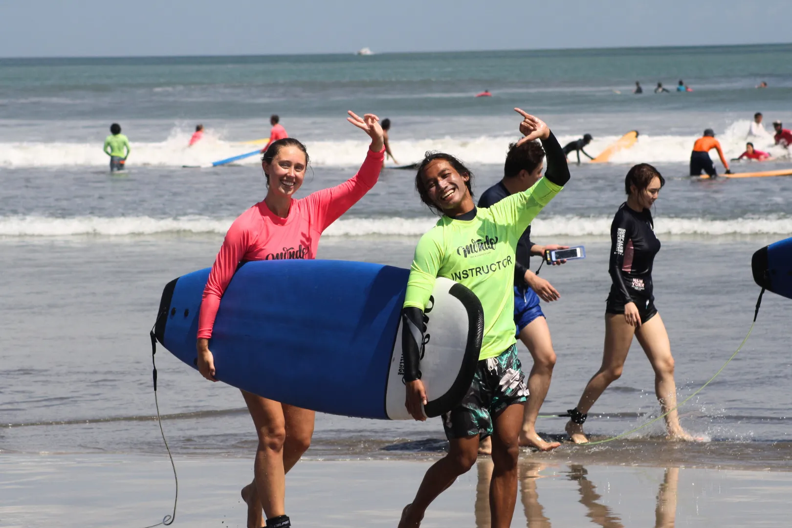 A smiling woman and a surf instructor carrying a blue surfboard together out of the ocean while making hand signs during a lesson at a surf camp Bali, with other surfers in the background.
