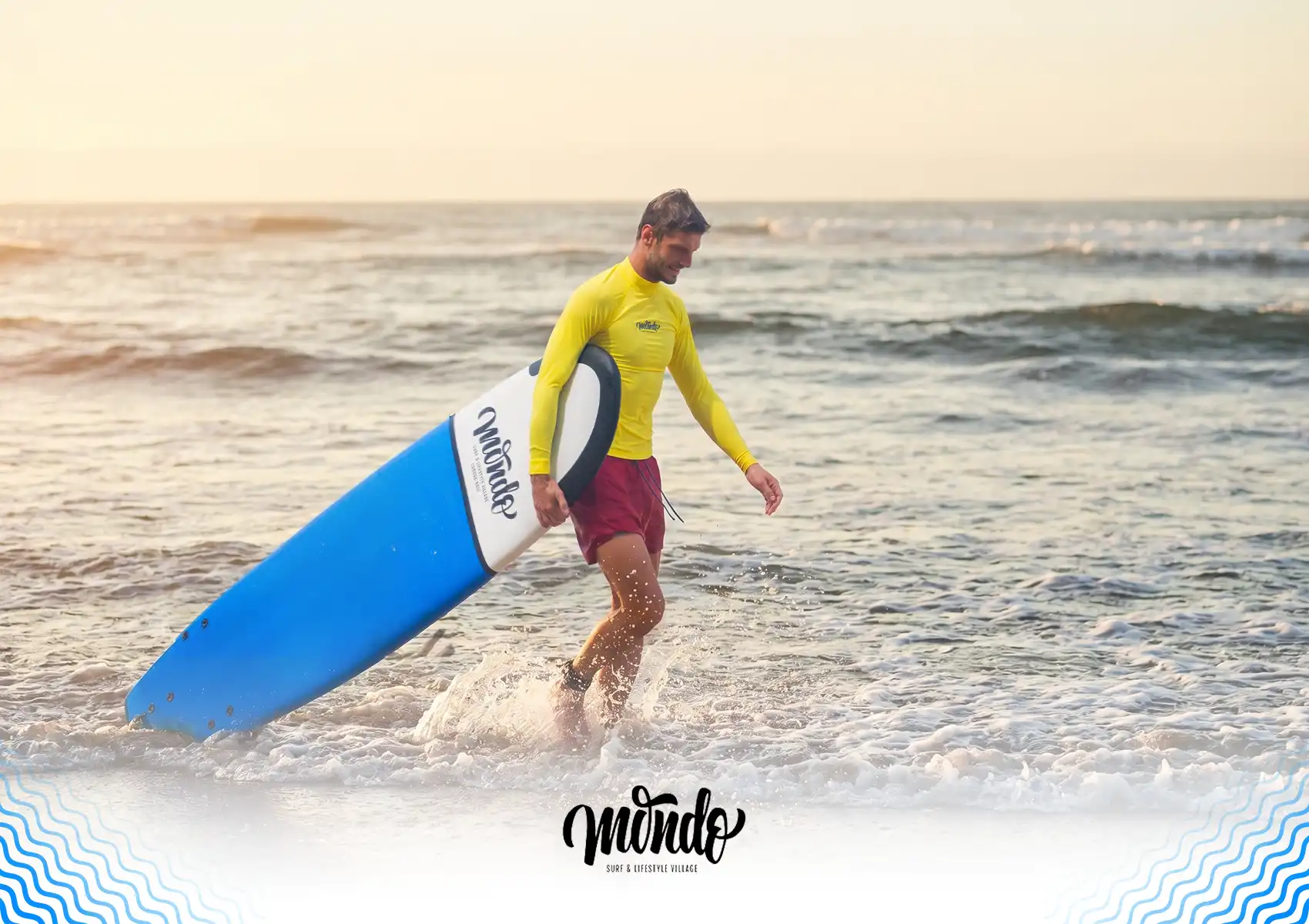 A smiling male surfer in a yellow rash guard walking out of the ocean carrying a blue surfboard at a surf camp, Bali.