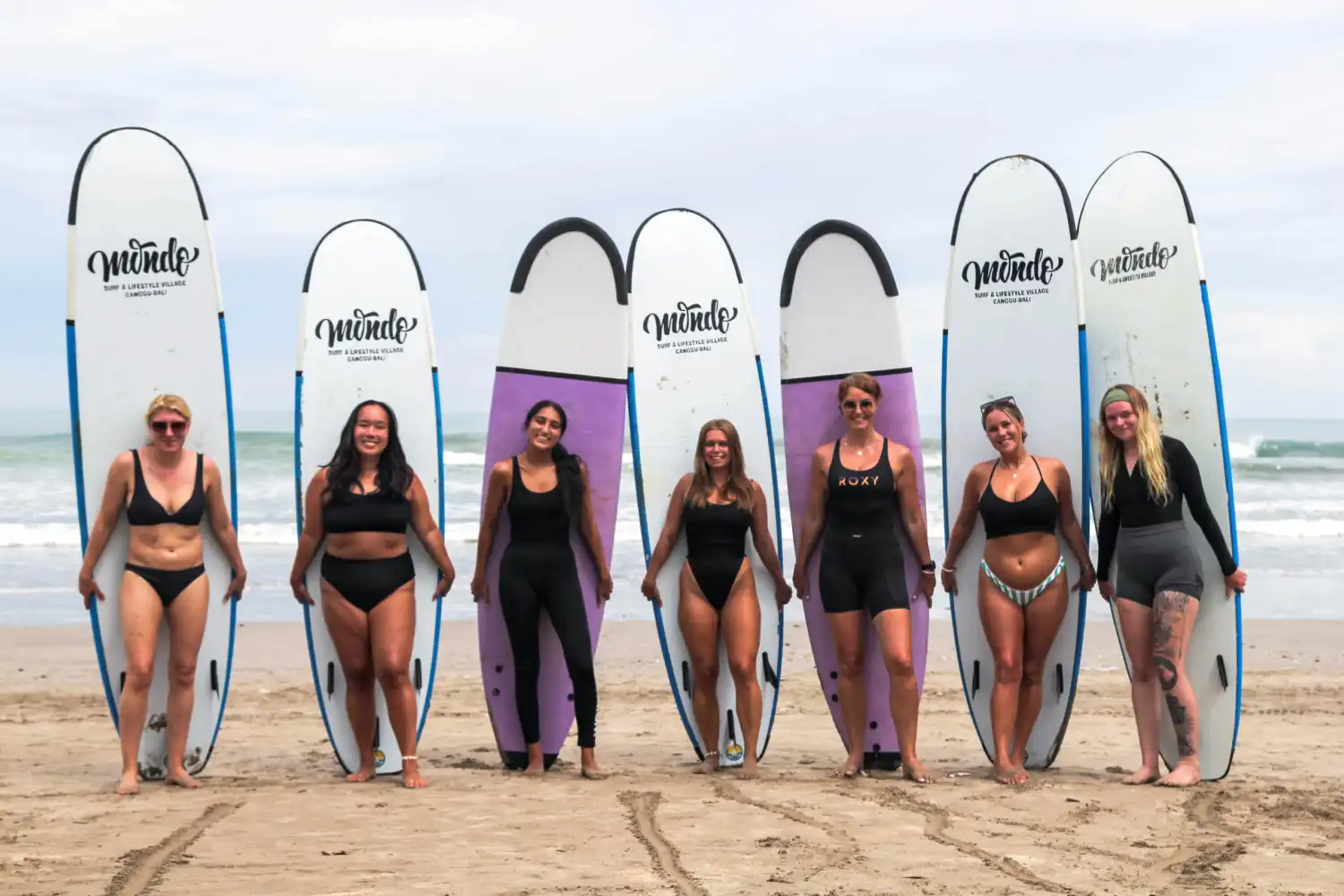 Seven smiling women standing in a row on a sandy beach, each holding a tall surfboard vertically beside them before a lesson at a surf camp Bali, with the ocean waves in the background