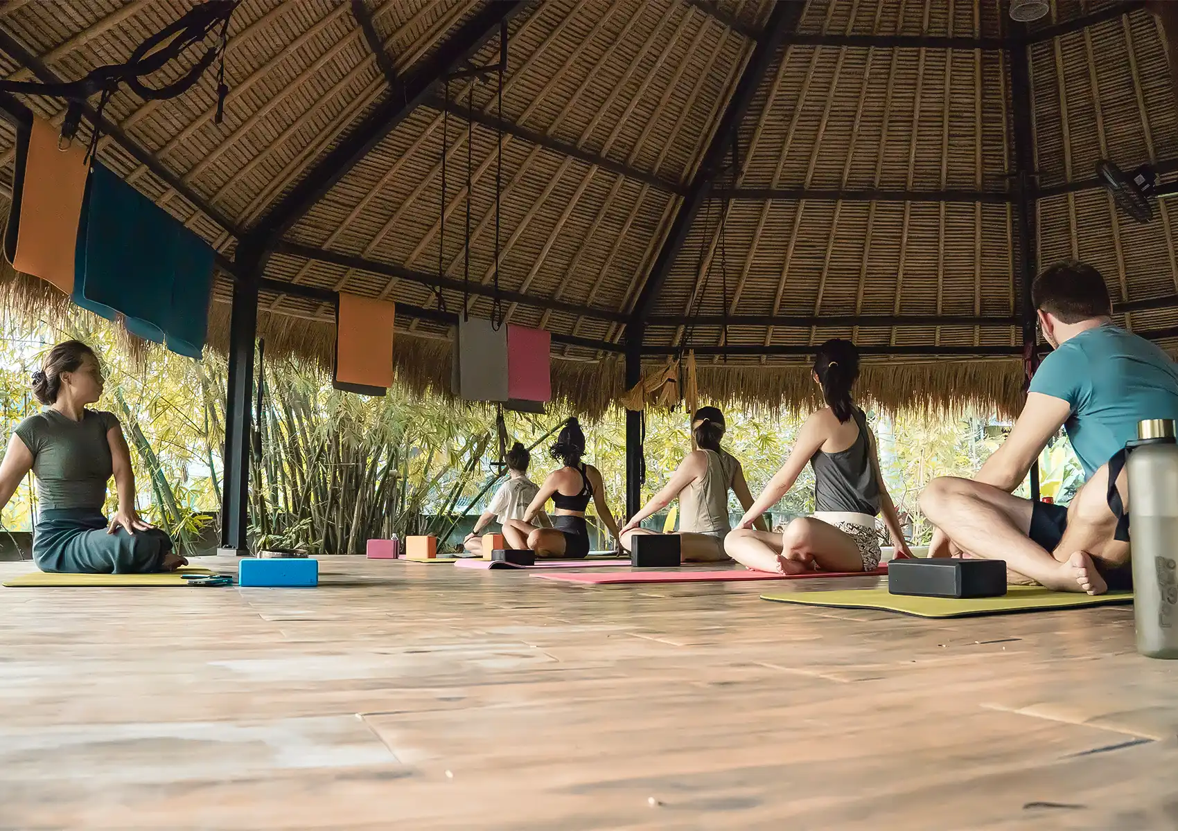 A group of people sit cross-legged on yoga mats during a class in an open-air thatched-roof studio surrounded by bamboo at Mondo Surf Village in Bali.