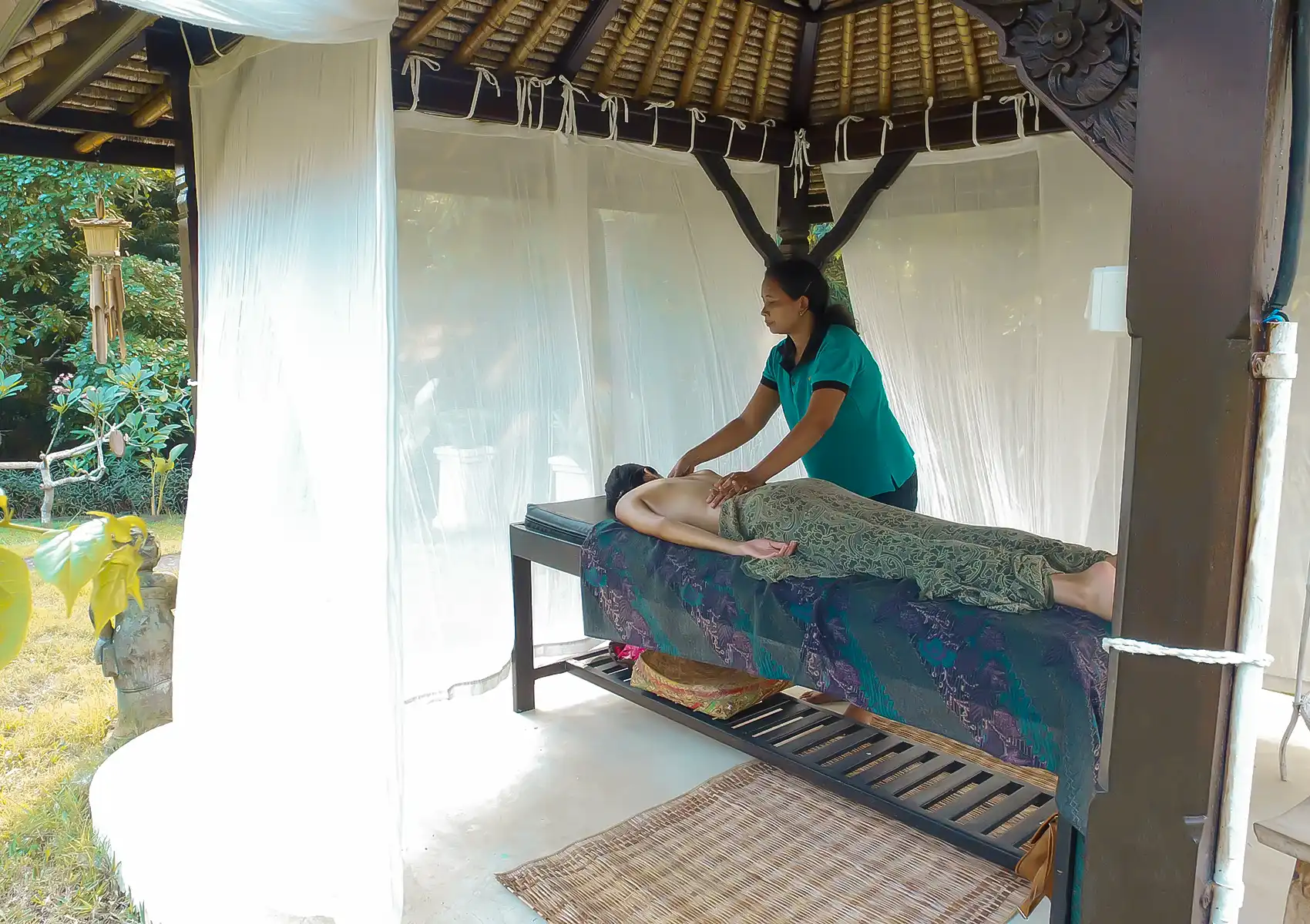 A therapist gives a massage to a person lying on a batik-draped table inside a breezy thatched-roof gazebo with mosquito netting, surrounded by tropical greenery at a surf village in Canggu, Bali.