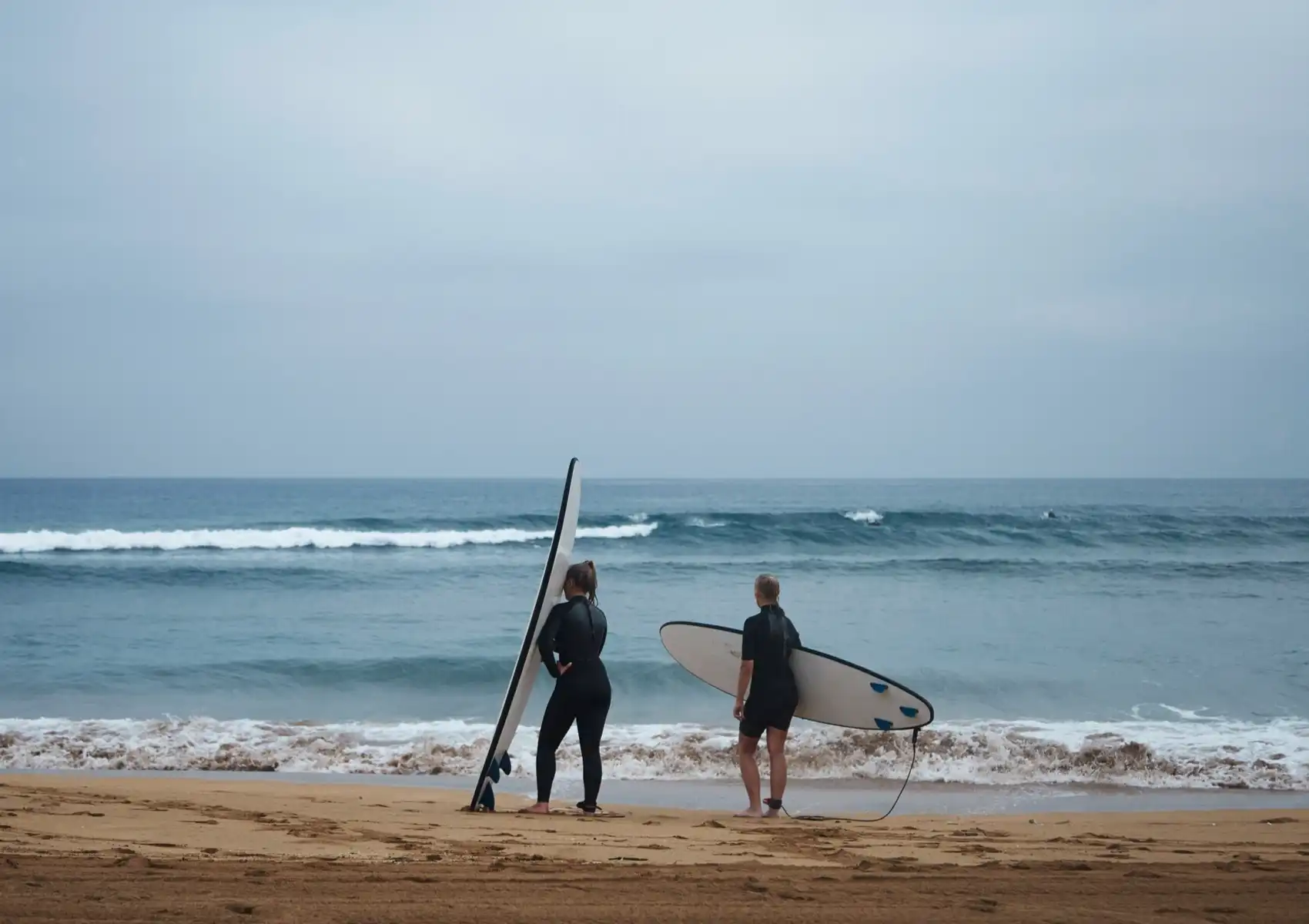 Two surfers in wetsuits stand on the beach holding their boards while studying the waves before paddling out at a surf camp in Bali.