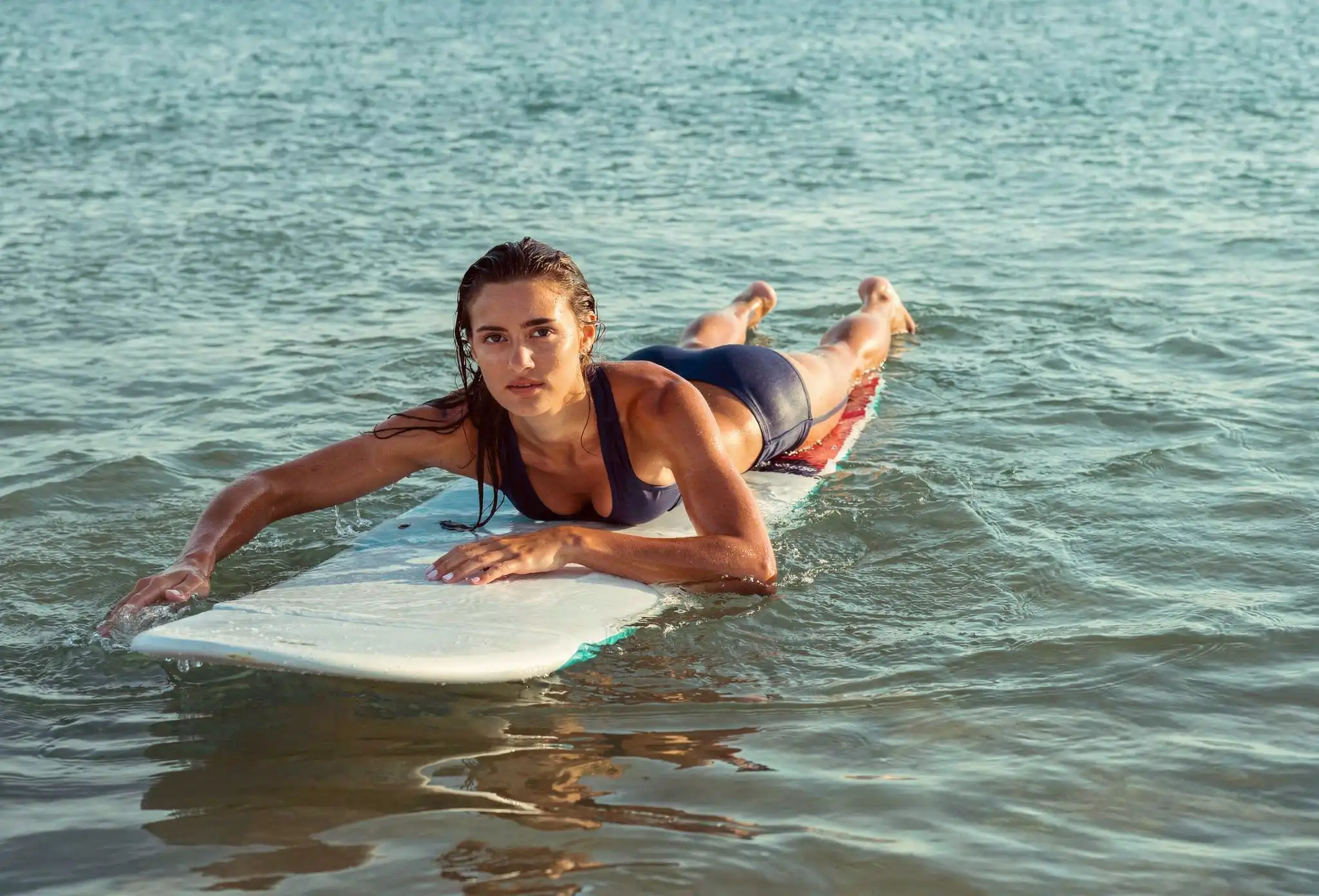 A woman in a navy bikini paddles efficiently on her surfboard through calm water at a surf camp in Bali.