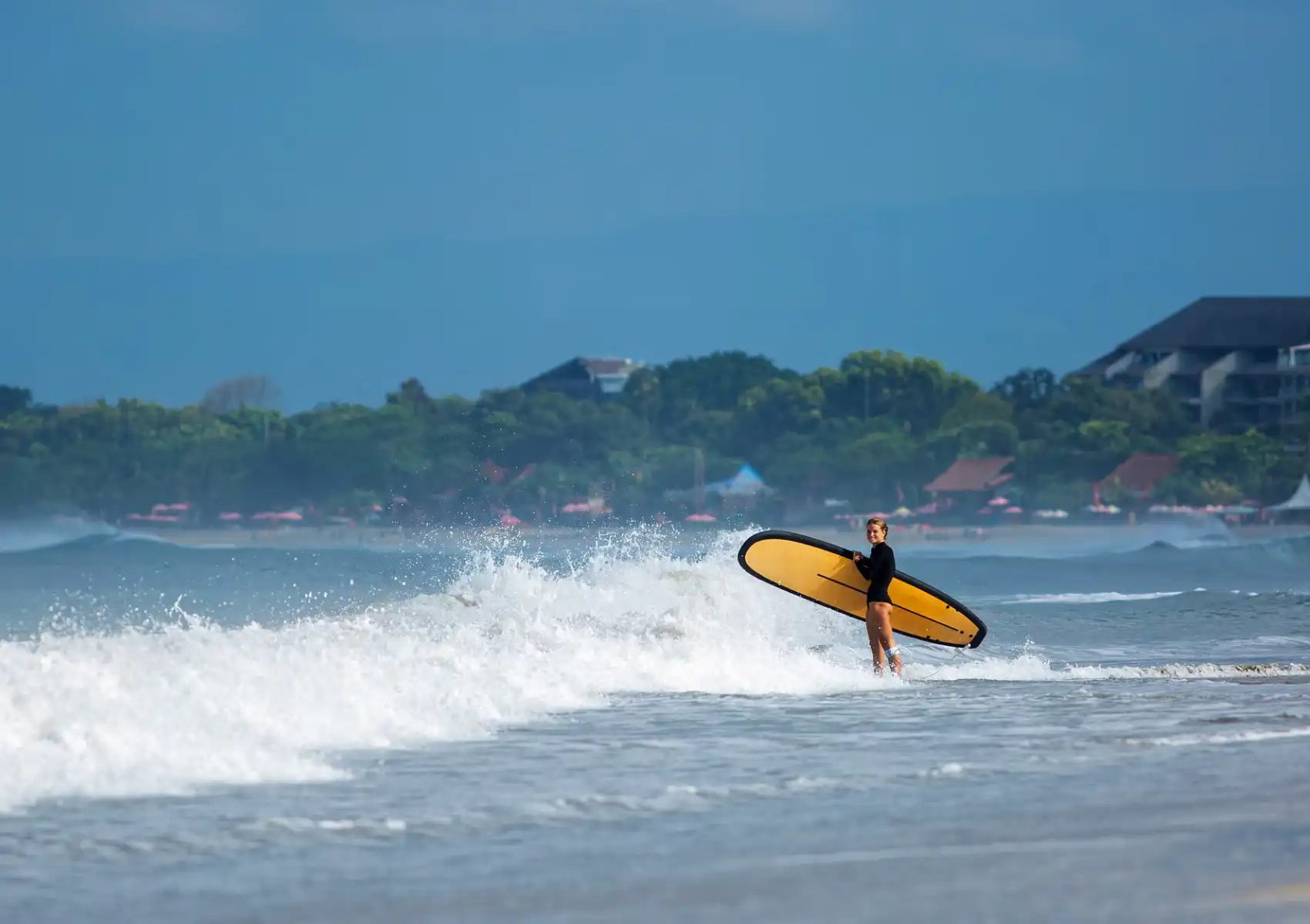A woman in a black wetsuit carries a yellow longboard through breaking whitewater toward shore, with a tree-lined coastline behind her at a surf camp in Bali.