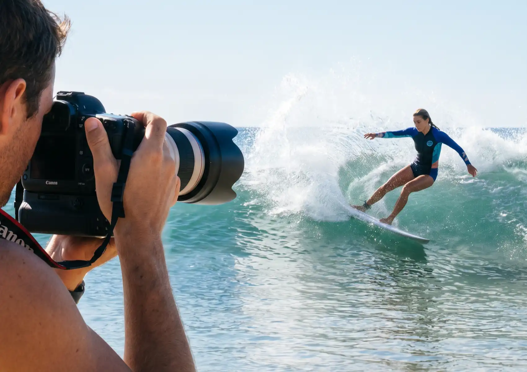 A photographer holds a Canon DSLR with a telephoto lens, shooting a woman in a blue wetsuit surfing on a breaking wave