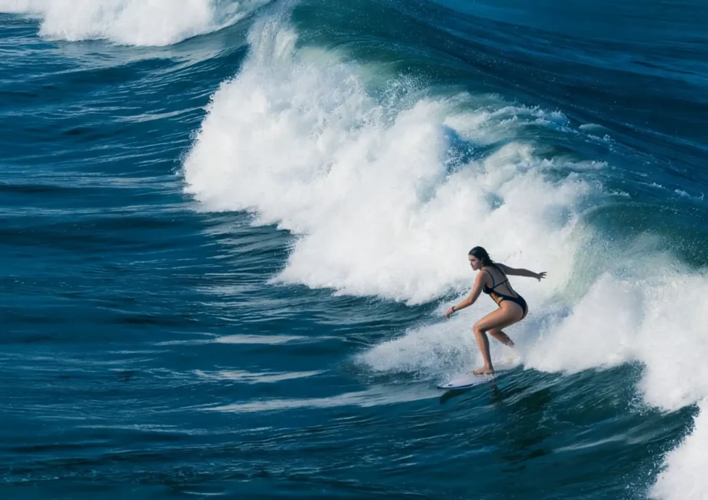 A woman in a black swimsuit rides the face of a powerful breaking wave over a reef break at a surf camp in Bali.
