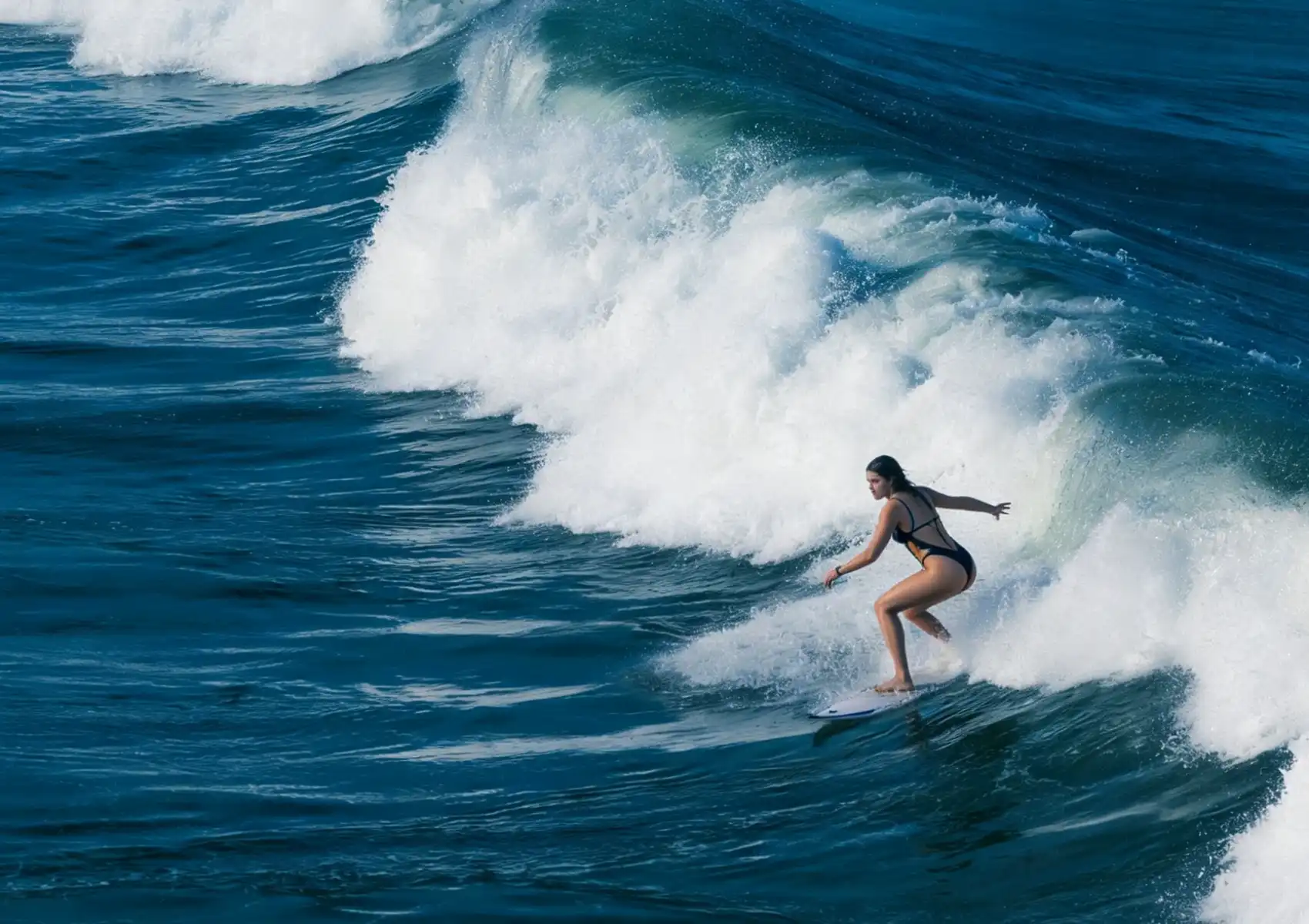 A woman in a black swimsuit rides the face of a powerful breaking wave over a reef break at a surf camp in Bali.