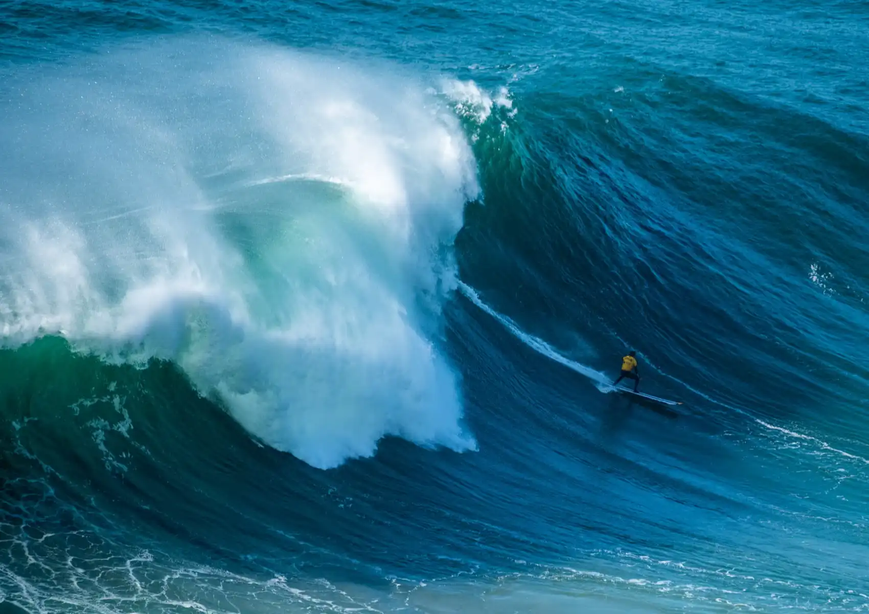 A surfer in a yellow top rides a thruster-finned board down the face of a huge powerful wave, showing the drive and control thruster fins offer at a surf camp in Bali.