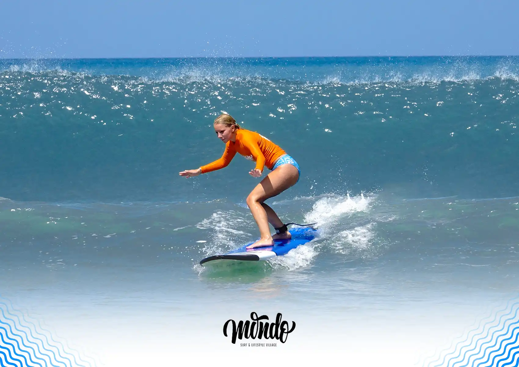 A female surfer wearing an orange rash guard balancing on a blue soft-top surfboard while successfully riding a wave at a surf camp bali.