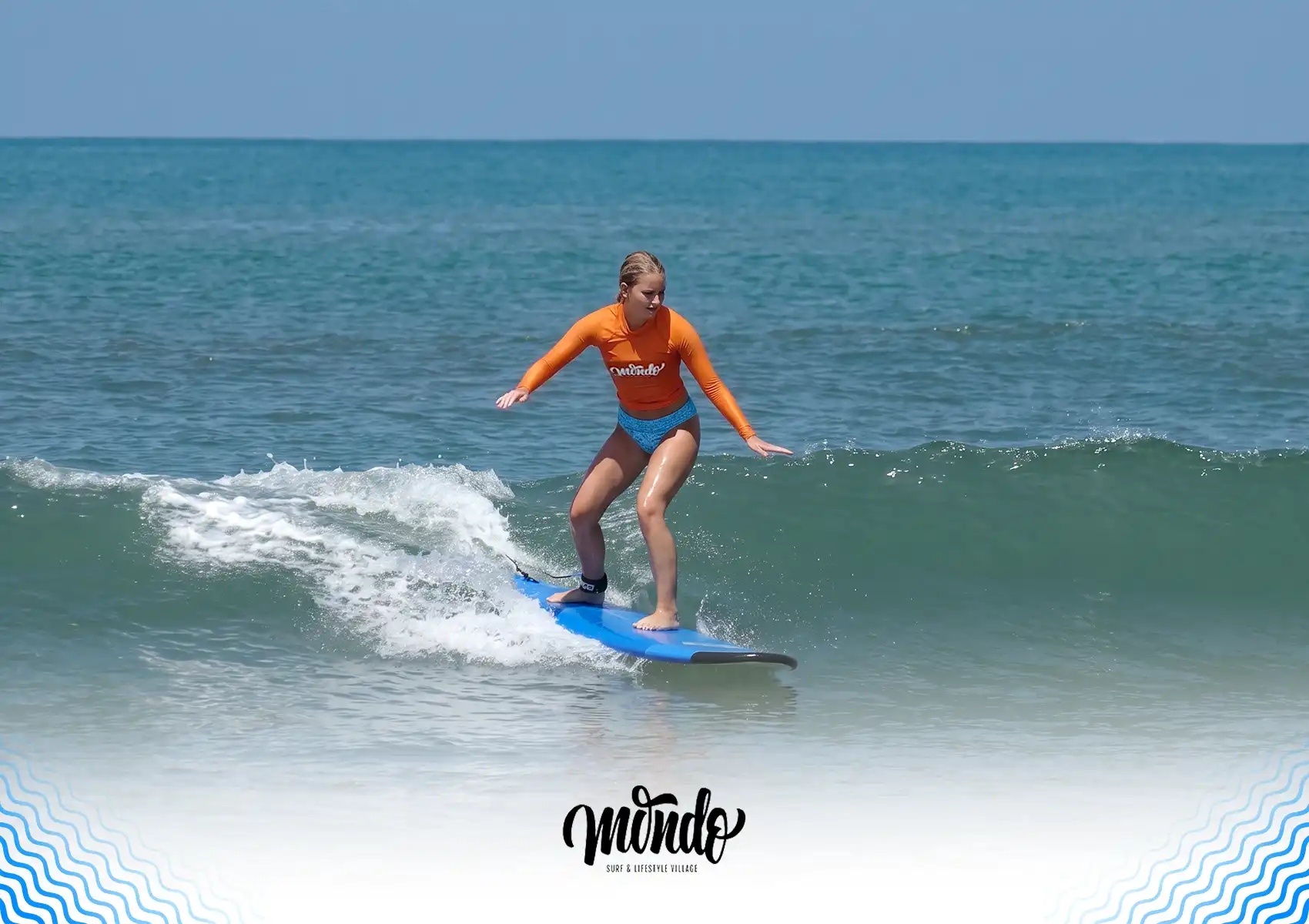 A female surfer in an orange rash guard demonstrating proper body position while successfully balancing on a blue surfboard on a small wave at a surf camp bali.