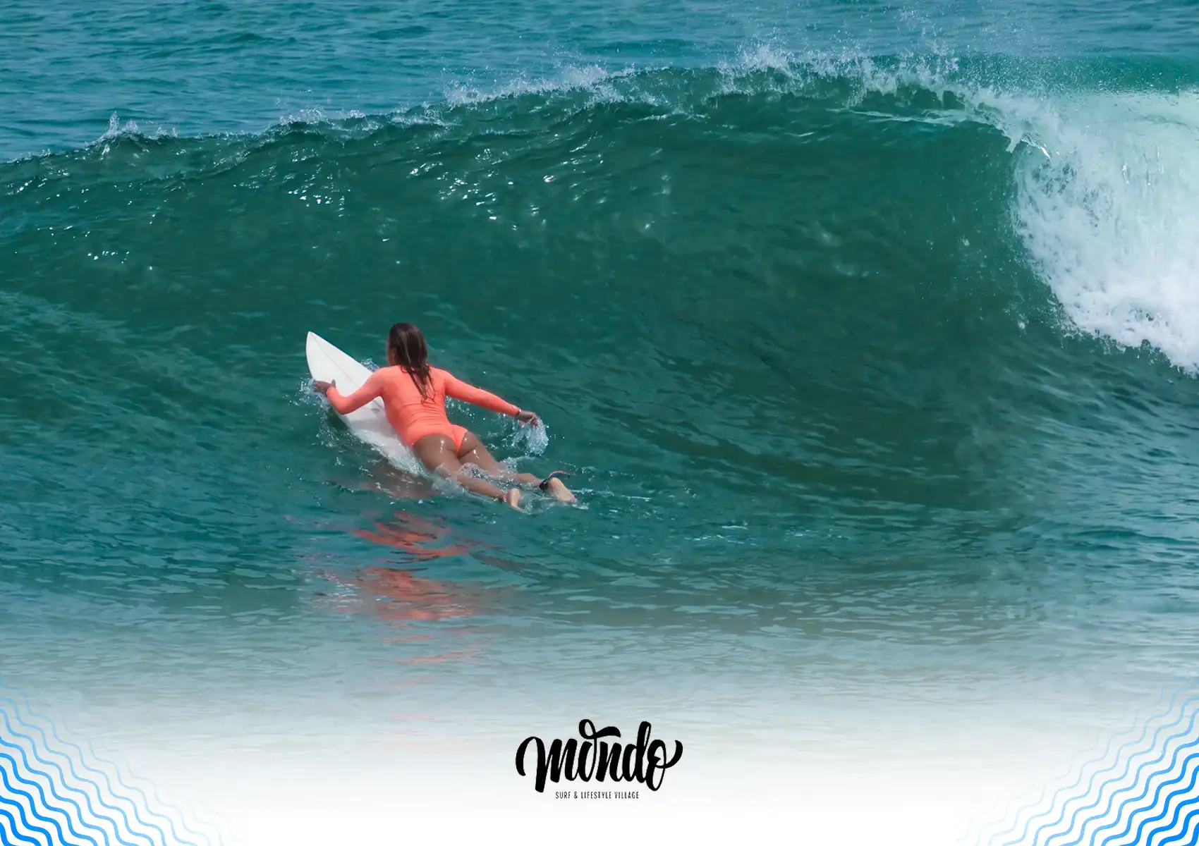 A female surfer in an orange swimsuit paddling a white surfboard up the face of a large, unbroken green wave during a session at a surf camp bali.