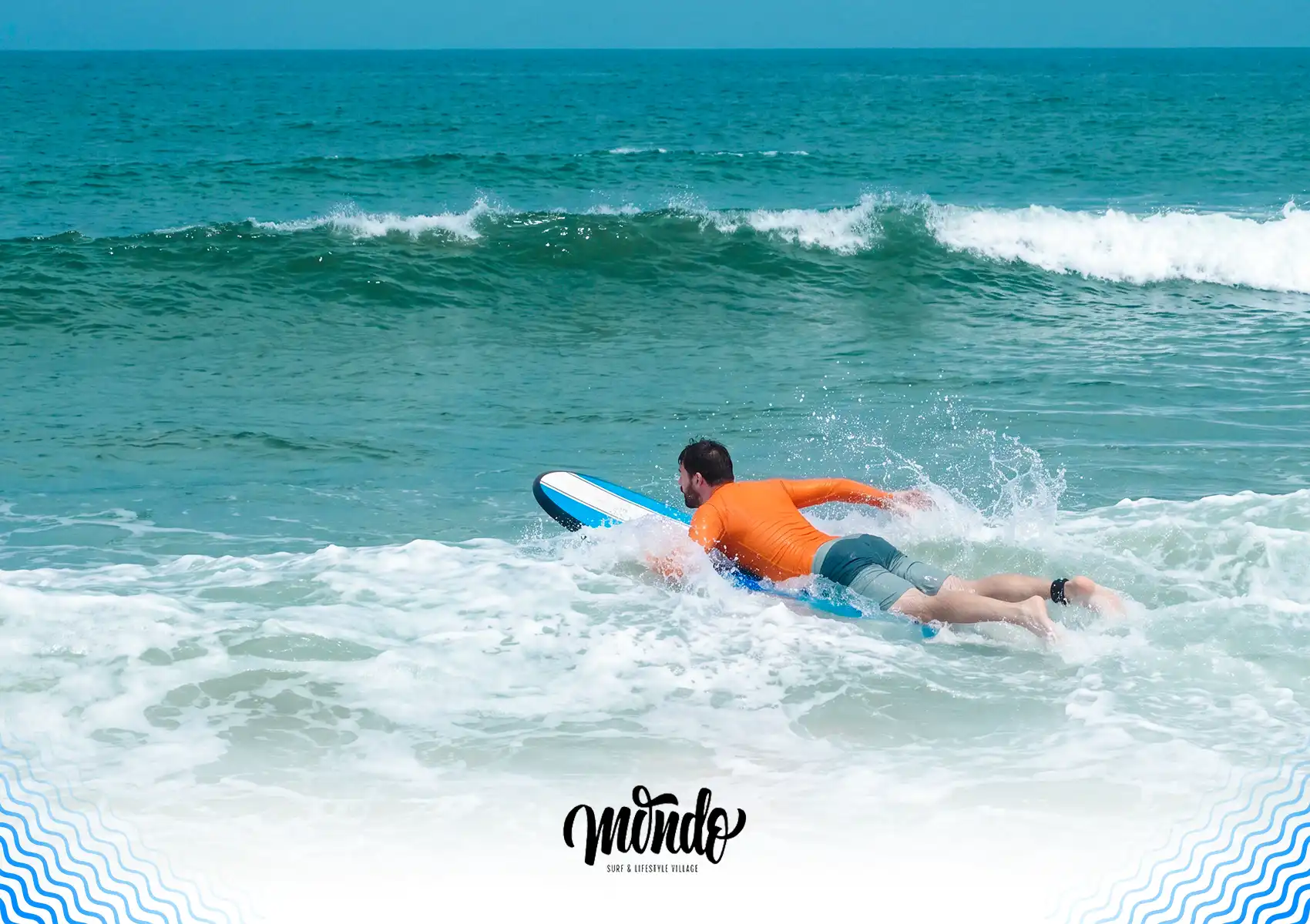 A male surfer in an orange rash guard paddling a surfboard through the whitewater to get past breaking waves at a surf camp bali.