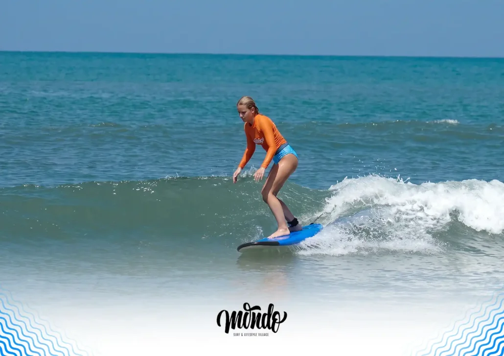 A female surfer in an orange rash guard shifting her weight and adjusting her stance to execute a turn on her blue surfboard while riding a gentle wave at a surf camp bali.