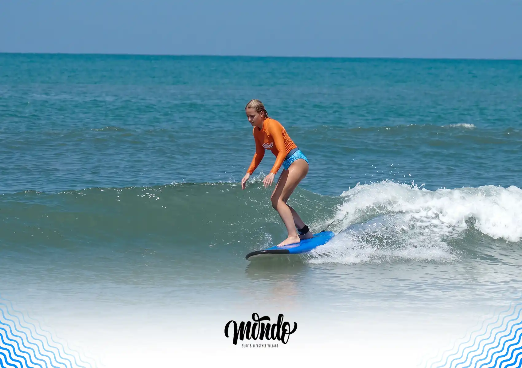 A female surfer in an orange rash guard shifting her weight and adjusting her stance to execute a turn on her blue surfboard while riding a gentle wave at a surf camp bali.