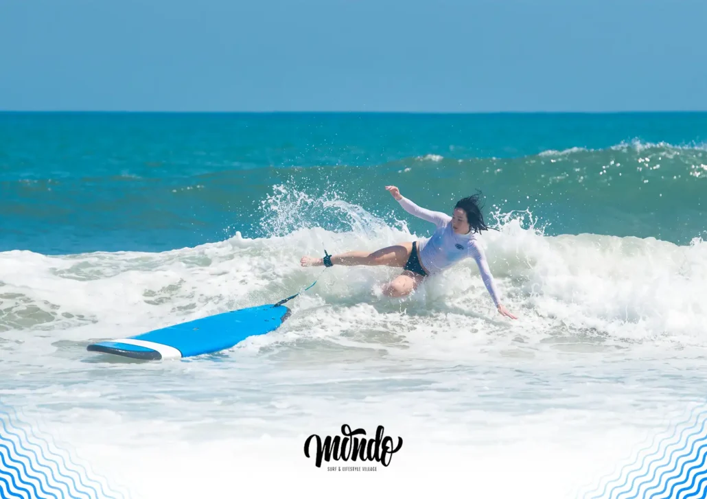 A female surfer in a white rash guard safely falling backwards off her blue surfboard into the ocean waves during a session at a surf camp bali.