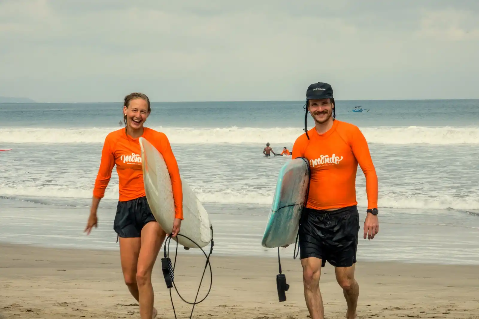 Two smiling surfers in orange rash guards carrying their boards on the beach at Mondo surf camp Bali.