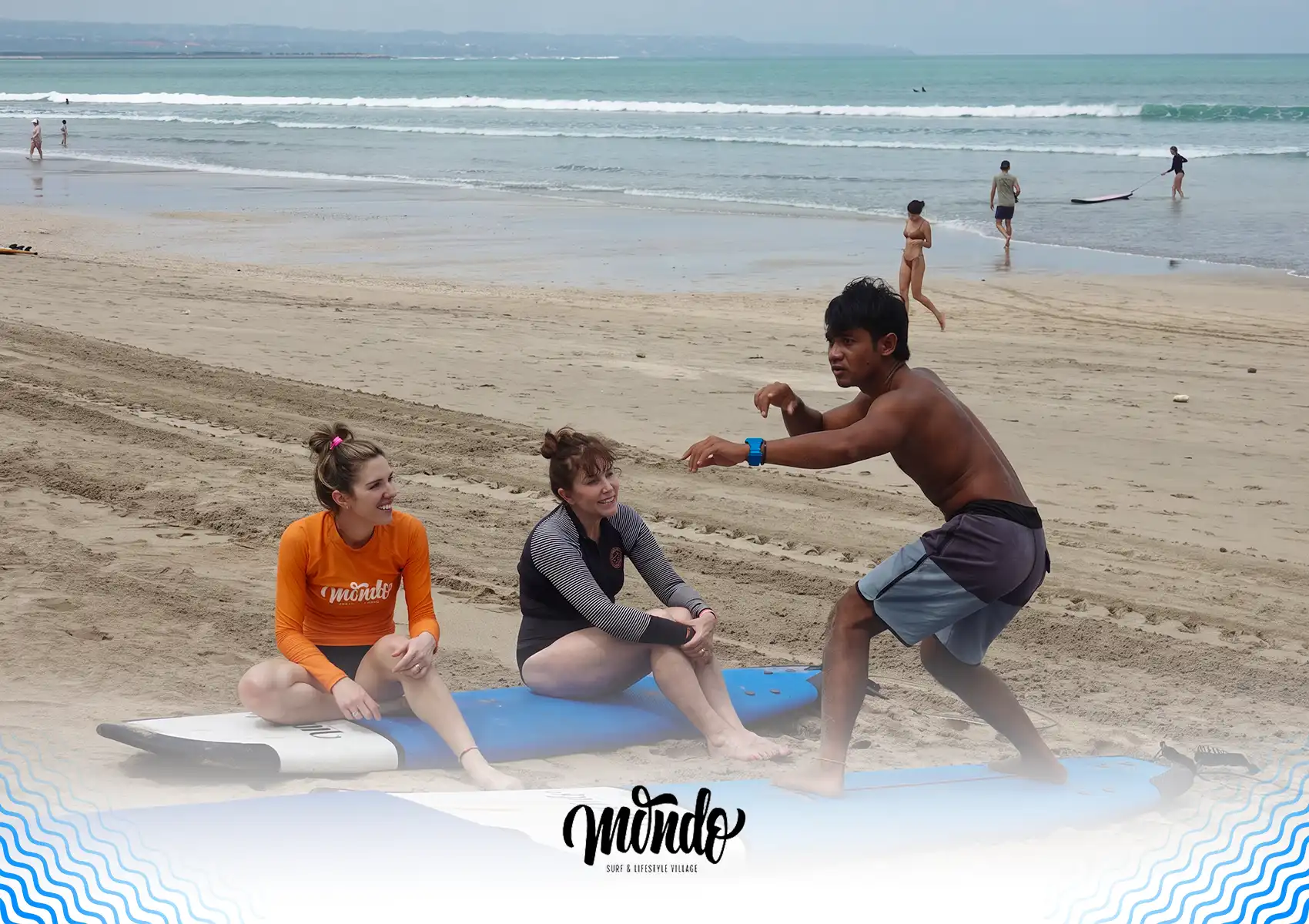 A surf instructor on a sandy beach demonstrating a low, balanced stance on a surfboard to two seated female students during a lesson at a surf camp bali.