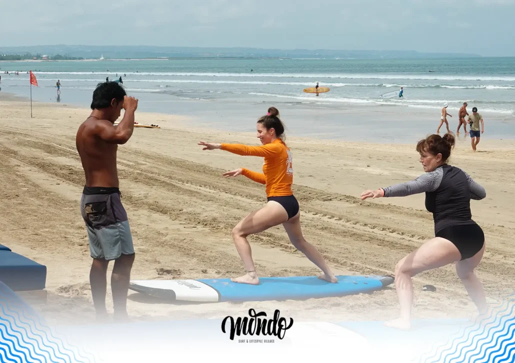 A surf instructor teaching two women the proper surfing stance as they practice popping up on their boards on the sandy beach during a lesson at a surf camp bali.