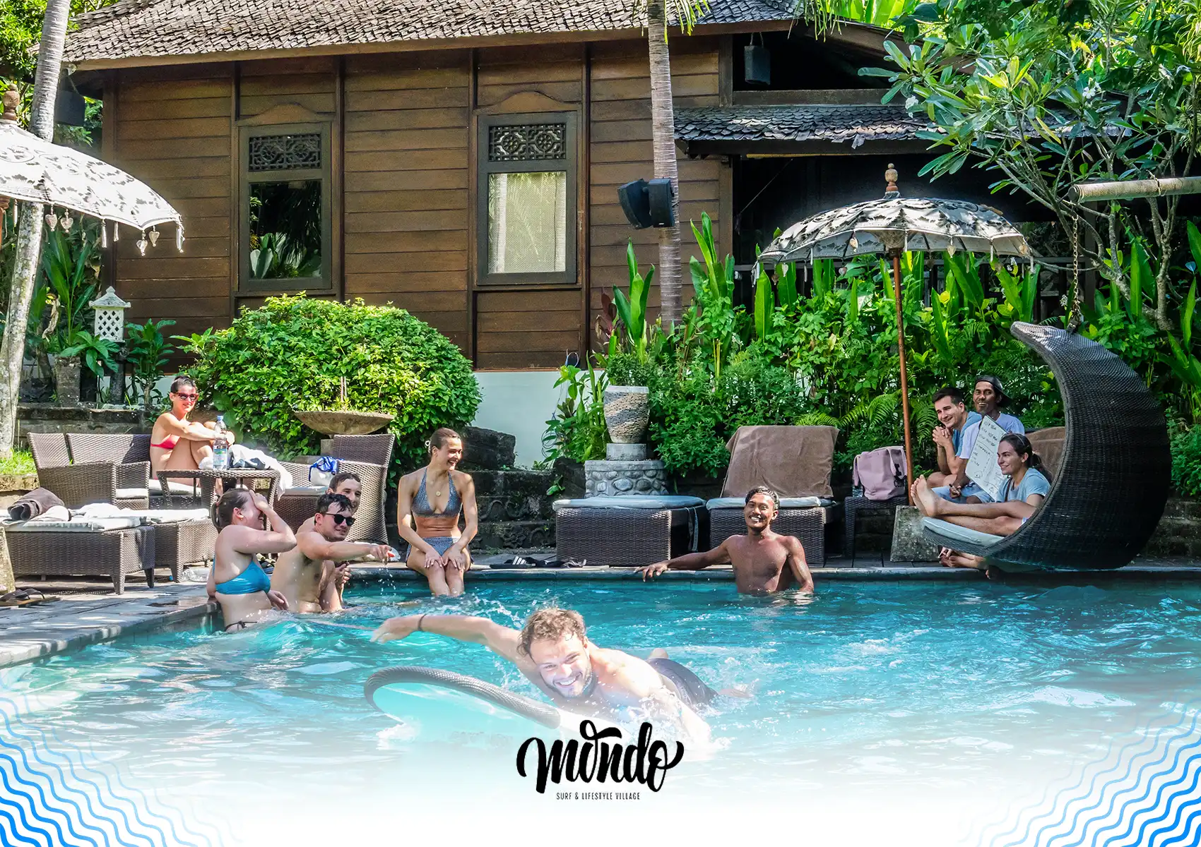A group of guests relaxing and socializing by a lush outdoor pool while a man practices paddling on a surfboard at a surf camp Bali.