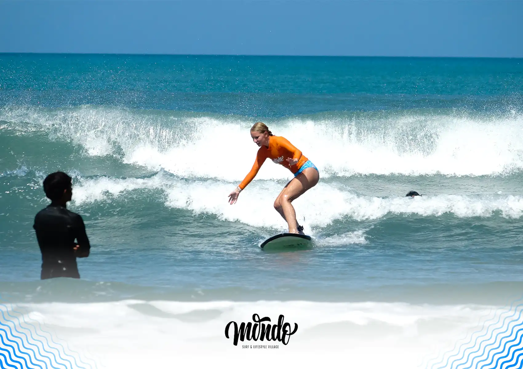 A female student in an orange rash guard successfully balancing on a surfboard while riding a wave, with her instructor watching closely from the water at a surf camp bali.