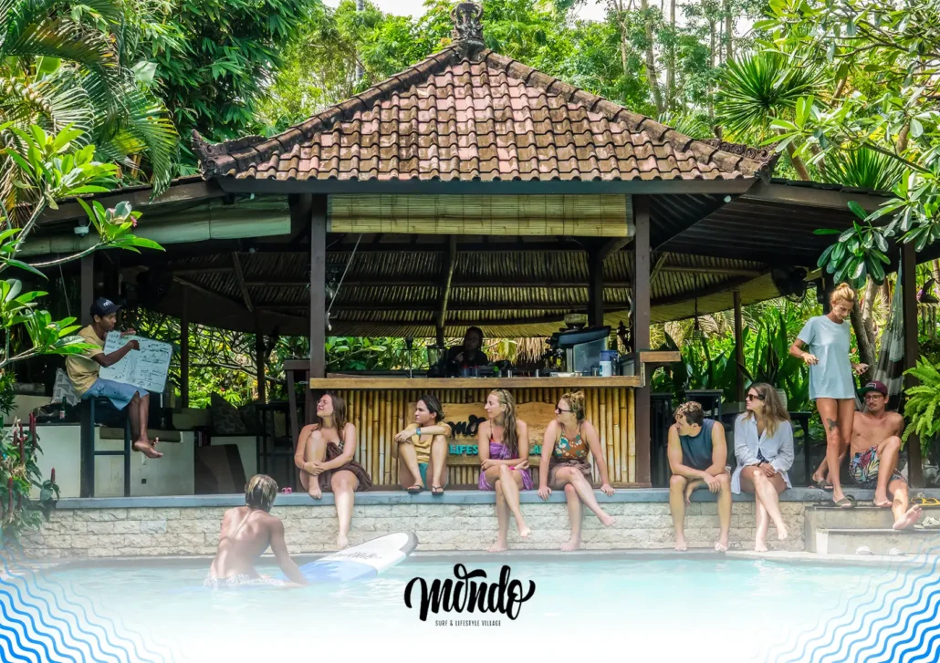 An instructor using a whiteboard to teach a surf theory lesson to a group of guests gathered along the edge of a swimming pool at a surf camp Bali