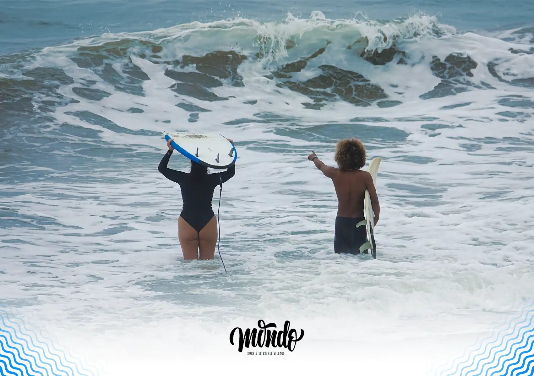 Two people standing waist-deep in the ocean holding their surfboards, with one pointing toward a large incoming wave during a session at a surf camp Bali.
