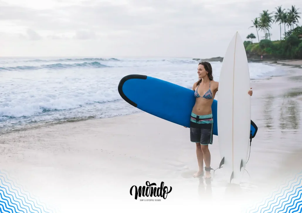 A female surfer standing on the sandy beach looking toward the ocean waves, holding a blue foam surfboard under one arm and a white hardboard upright in the other at a surf camp Bali.