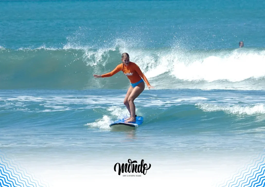 A woman in an orange rash guard successfully balancing on her blue surfboard while riding a small wave at a surf camp Bali.