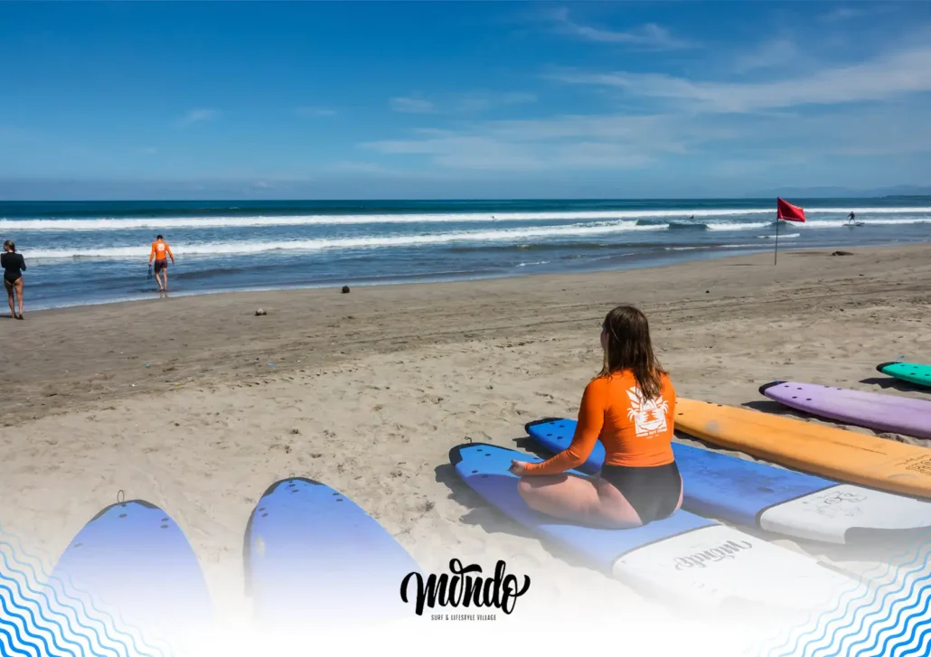 A woman wearing an orange rash guard sitting cross-legged on her surfboard on the sandy beach, observing the ocean waves before a lesson at a surf camp Bali.