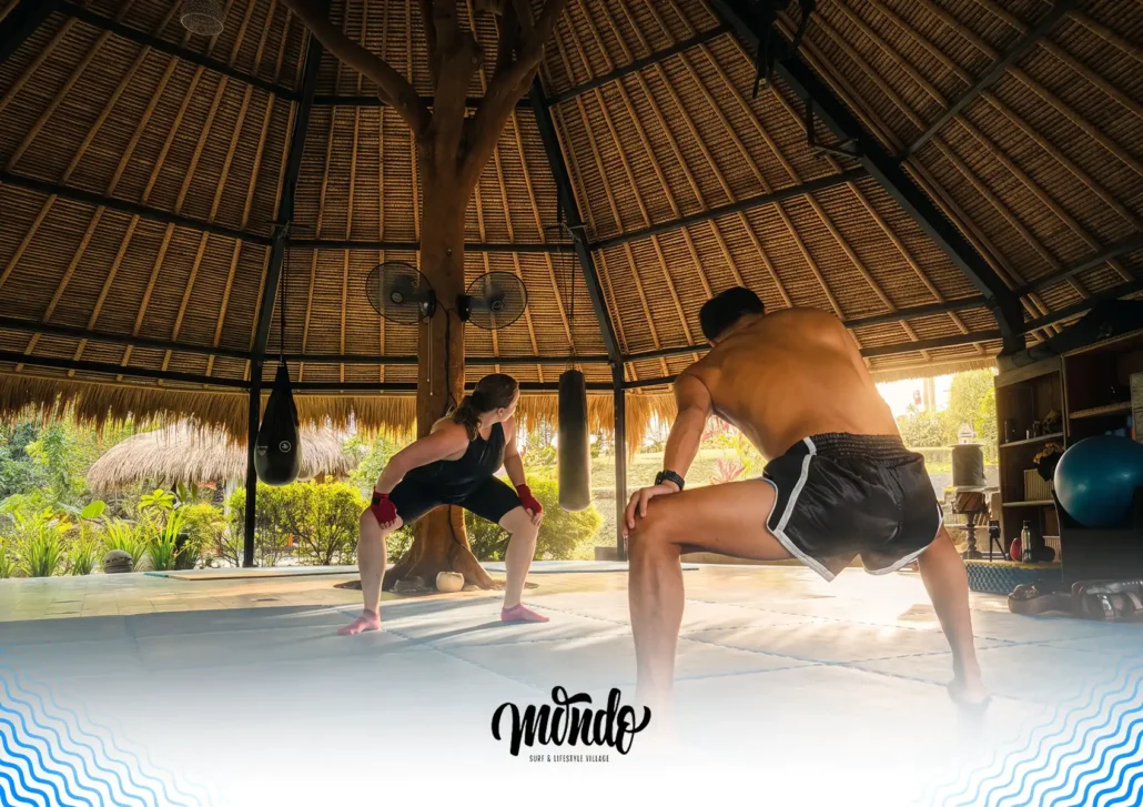 A woman and a male instructor performing a wide-legged squat stretch on training mats inside a spacious thatched-roof studio built around a large central tree at a surf camp Bali.