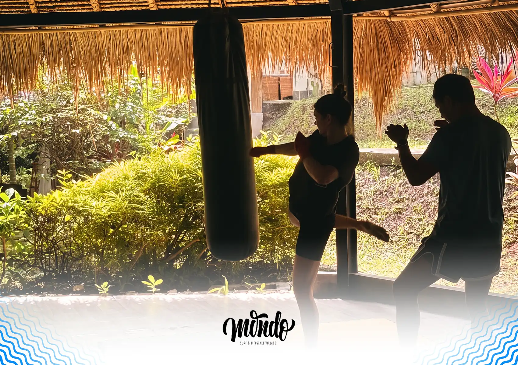 A woman practicing a knee strike on a heavy punching bag while an instructor guides her inside an open-air, thatched-roof martial arts studio surrounded by tropical plants at a surf camp Bali.