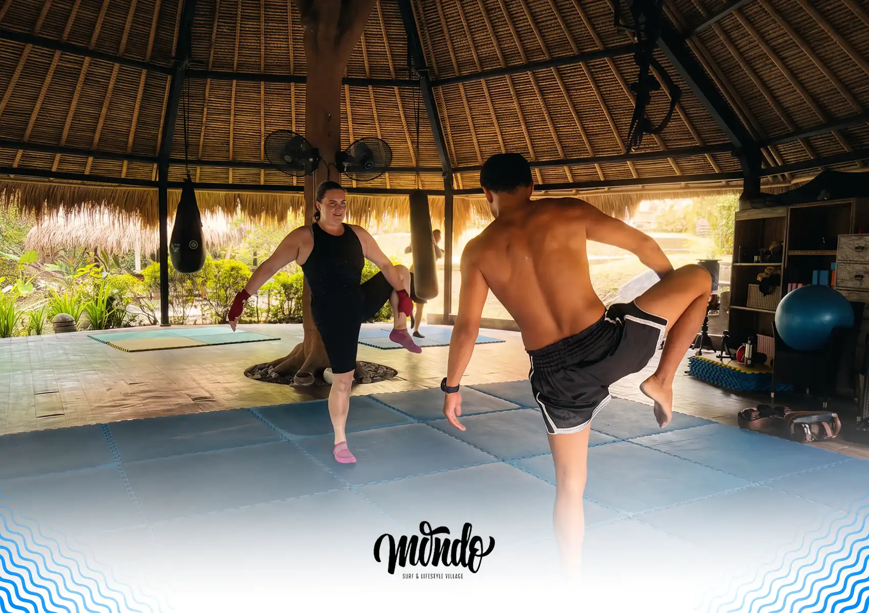 A woman and a male instructor performing a standing leg warm-up on blue training mats inside a tropical, open-air studio at a surf camp Bali.