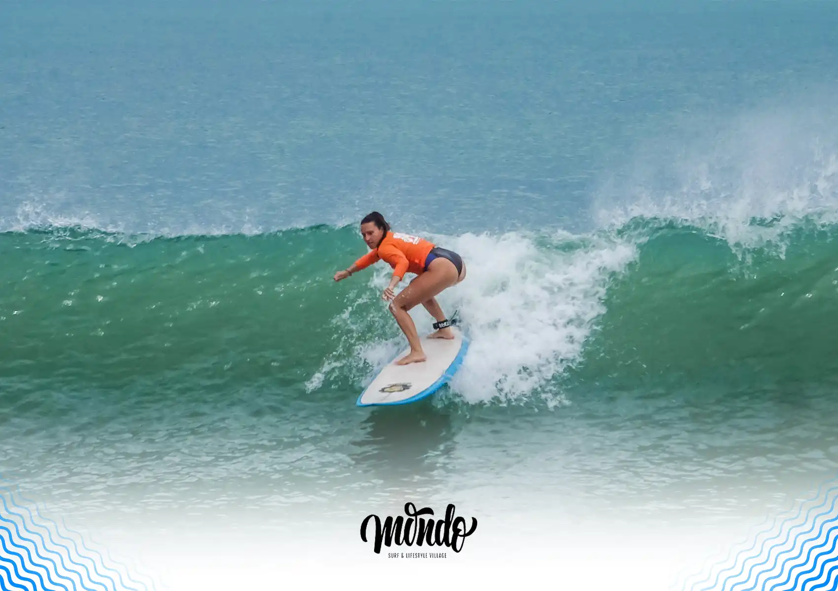 A woman wearing an orange rash guard riding a blue surfboard on a curling green wave at a surf camp Bali.