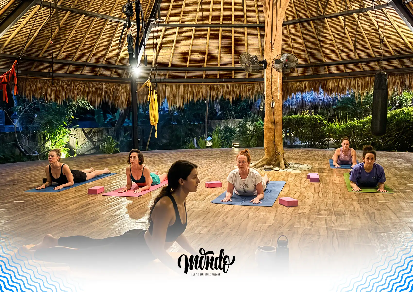 A group of women practicing the Sphinx yoga pose on mats in a spacious open-air studio during an evening session at a surf camp Bali.