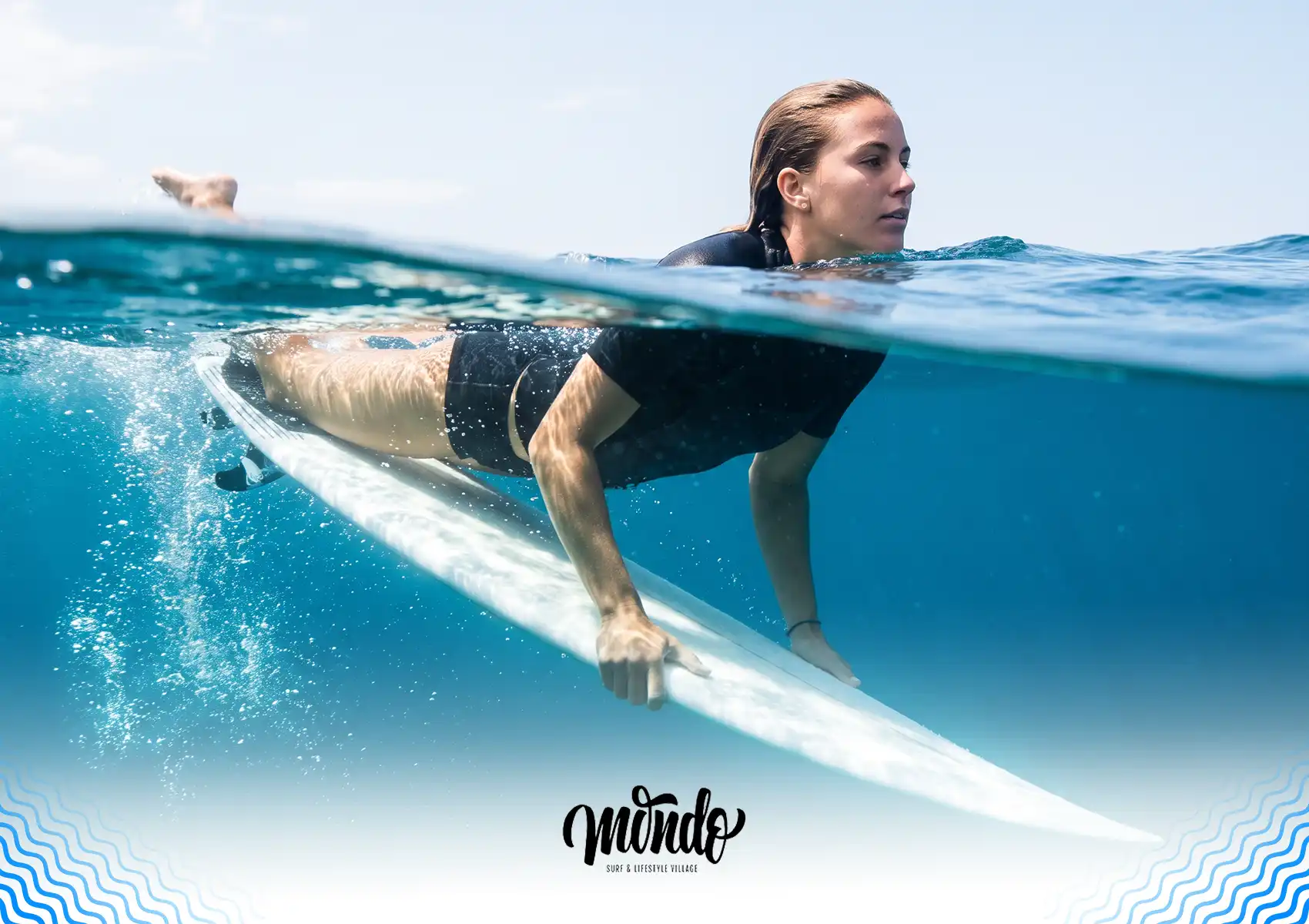 A split-level surface and underwater shot of a female surfer in a black wetsuit pushing the nose of her surfboard down to initiate a duck dive while training at a surf camp bali.